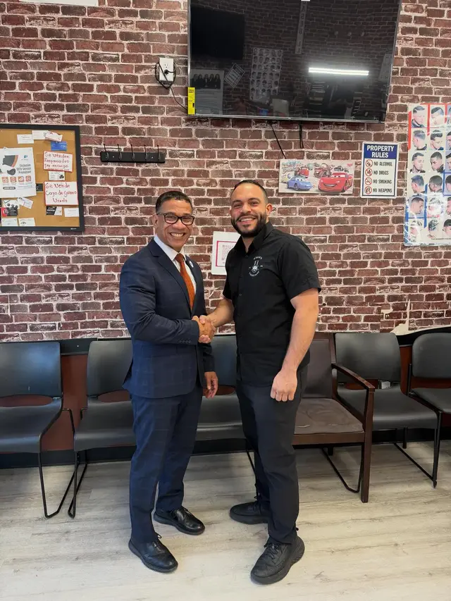 Two men shaking hands and smiling inside Mago's Barbershop, one in a navy suit and one in a black Mago's Barbershop shirt